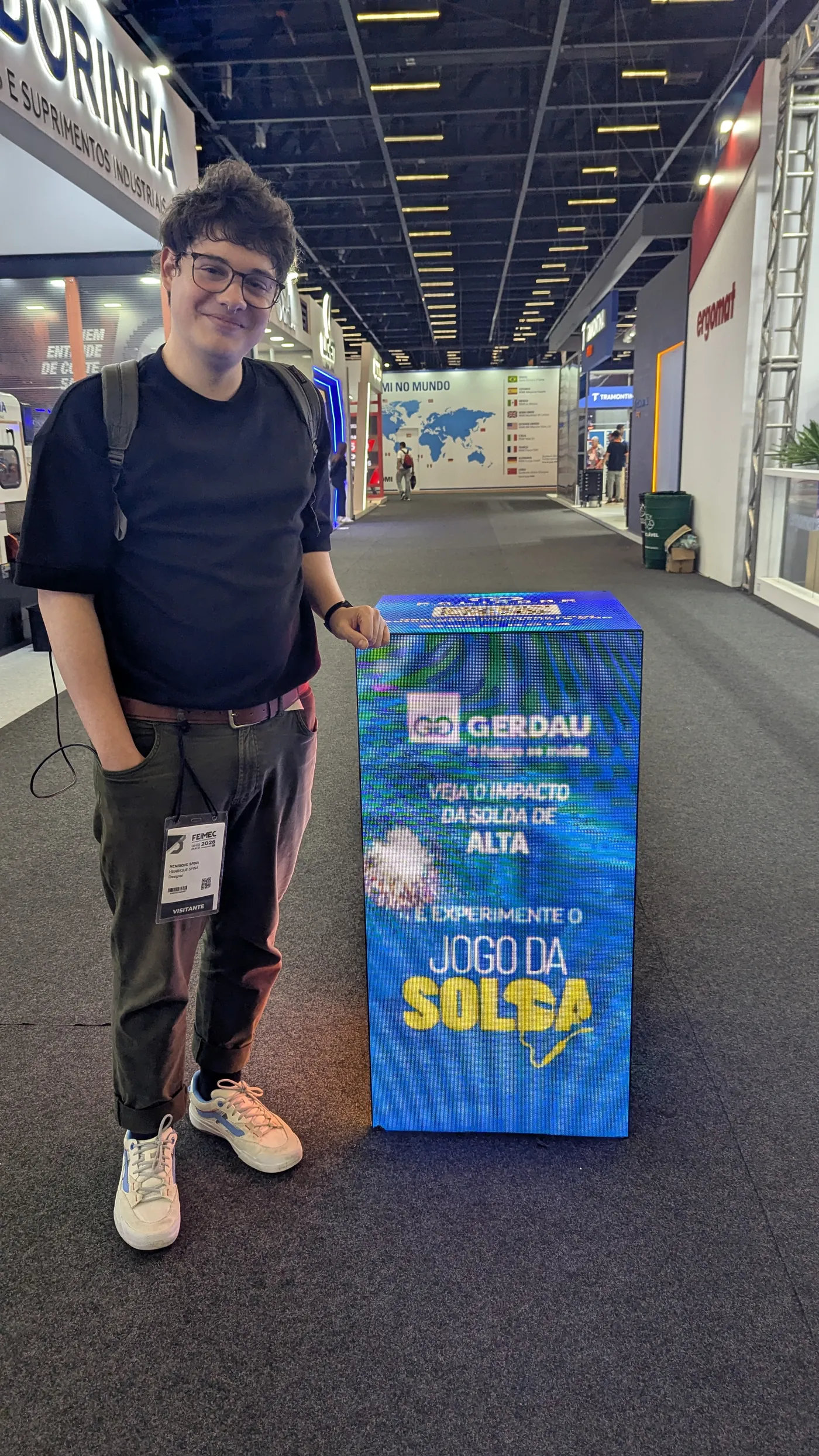 Henrique ao lado do totem azul Jogo da Solda no corredor da FEIMEC